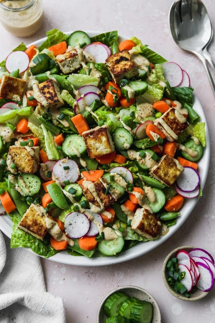 Overhead photo of a large white plate topped with a colourful chopped salad topped with tofu cubes and a drizzle of tahini dressing.