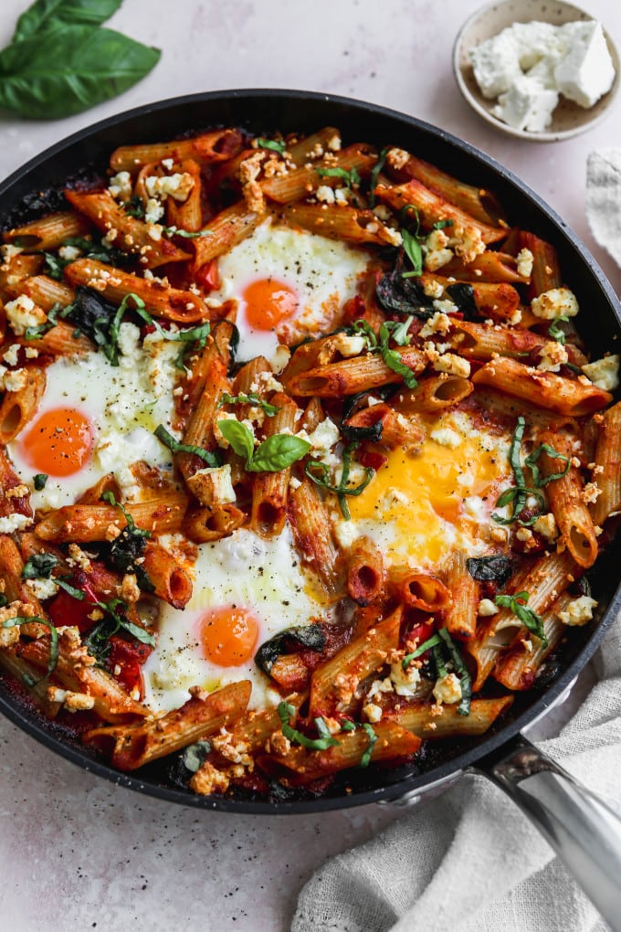 Side angle photo of a baked tomato pasta with eggs, feta, and basil in a black skillet.