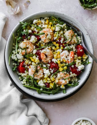 Overhead photo of arugula, couscous, roasted corn, tomatoes, and shrimp in a white bowl.
