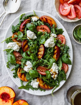 Overhead photo of a grilled nectarine, arugula, tomato, and burrata salad on a white serving dish.