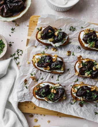 Overhead photo of crostini topped with whipped goat cheese and stewed prunes on a wood cutting board.