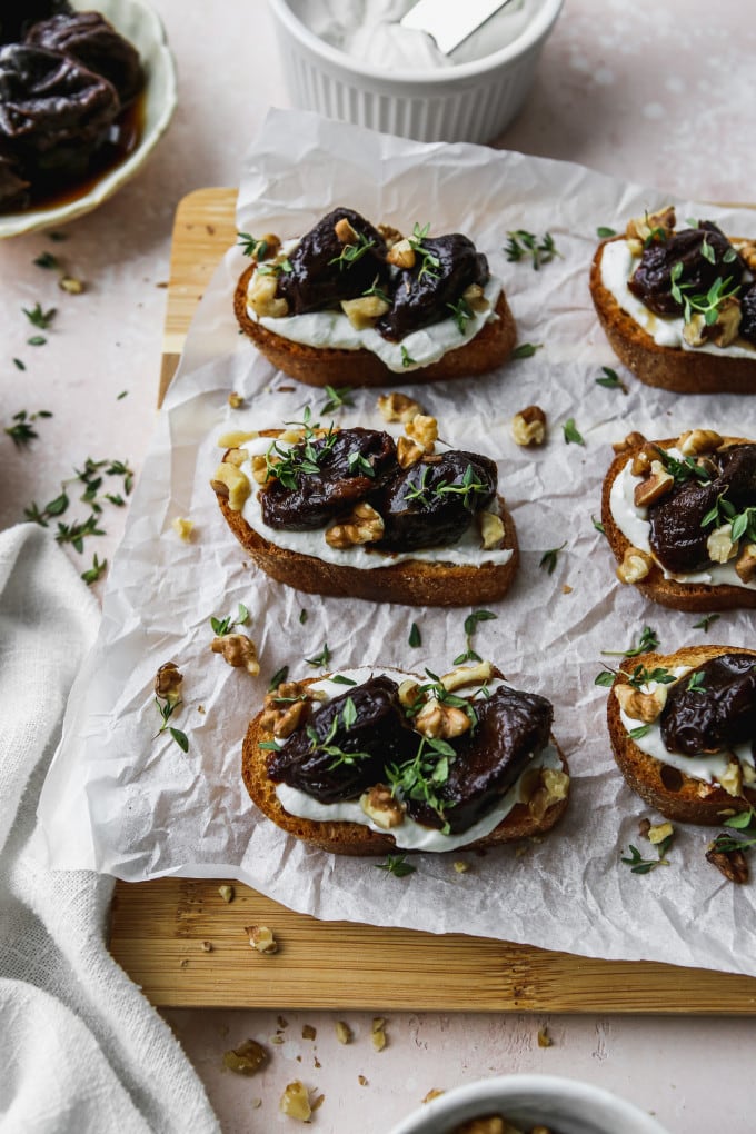 Overhead photo of crostini topped with whipped goat cheese and stewed prunes on a wood cutting board.