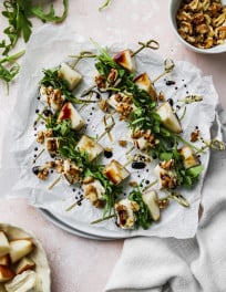 Overhead photo of pear, brie, arugula, and walnut salad skewers with a balsamic glaze on a white plate.