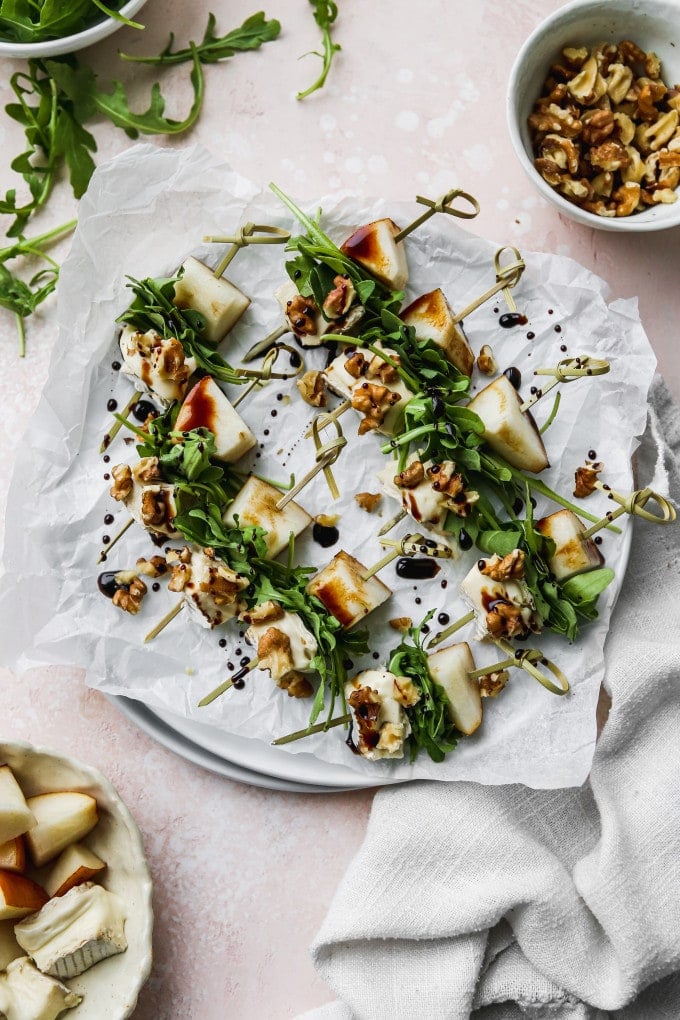 Overhead photo of pear, brie, arugula, and walnut salad skewers with a balsamic glaze on a white plate.