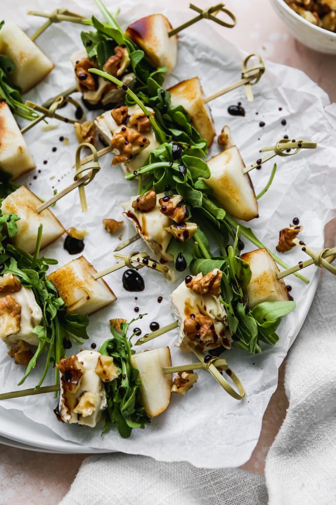 Closeup photo of pear, brie, arugula, and walnut salad skewers with a balsamic glaze on a white plate.