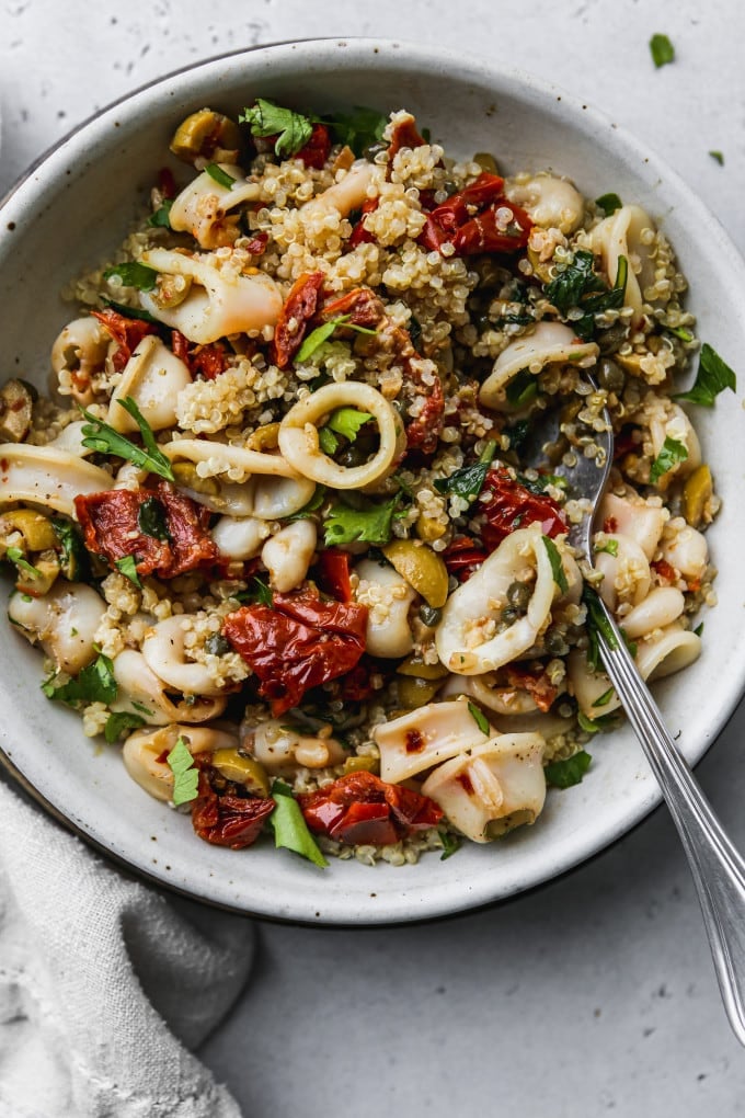 Overhead photo of sautéed squid rings, sun dried tomatoes, olives, and quinoa in a white bowl.