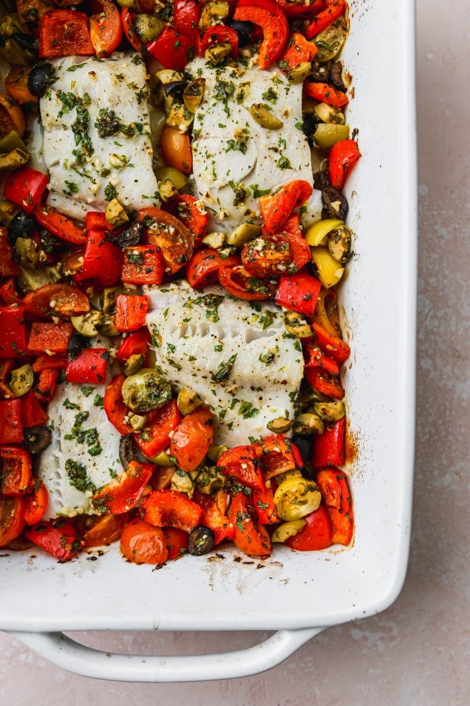 Baked black cod with tomatoes, peppers, and olives in a white baking dish.