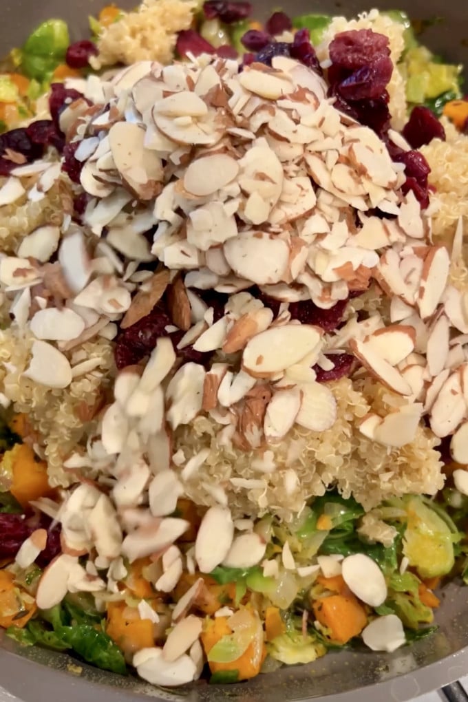 Adding cooked quinoa, dried cranberries, and sliced almonds to veggies in pan.